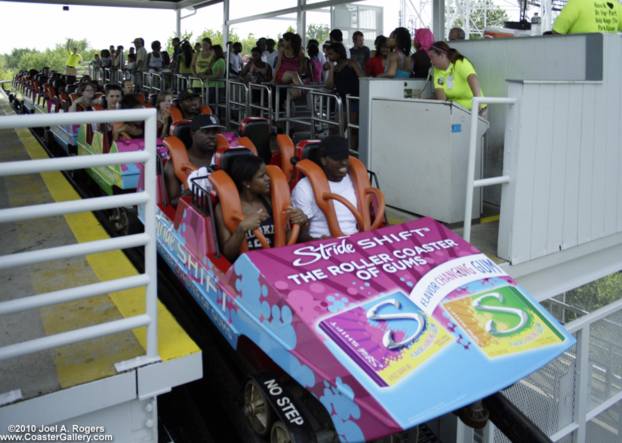 Rainbow colored roller coaster in the station. The train is currently wrapped in a gum advertisement